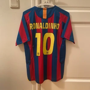 FC Barcelona Ronaldinho Retro - Klassisk FC Barcelona fotbollströja med Ronaldinho och nummer 10 på ryggen. Tröjan är randig i rött och blått med gula detaljer, klubbmärke på bröstet och Nike-logga. Tillverkad i lätt och ventilerande material, perfekt för dig som älskar fotboll och retrostil.