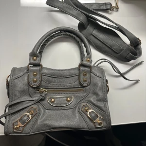 Grå handväska med gulddetaljer - Snygg grå handväska med guldfärgade metalldetaljer och dekorativa nitar. Väskan har både handtag och en avtagbar axelrem, samt en praktisk dragkedja framtill. Perfekt storlek för det viktigaste! Aldrig använd 👜