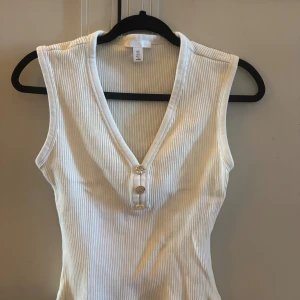 Vit ribbad linnetopp från H&M - Säljer en vit ribbad linnetopp från H&M med djup v-ringning och tre dekorativa knappar framtill. Toppen är ärmlös och har en figurnära passform. 