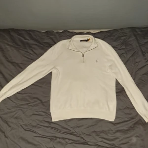 Vit half zip tröja från Polo Ralph Lauren - Vit långärmad tröja från Polo Ralph Lauren med half zip och klassisk krage. Tröjan har broderad logga på bröstet och är tillverkad i mjuk bomull. Perfekt för en clean och stilren look.
