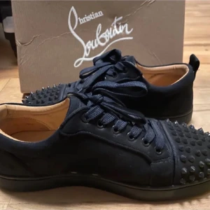 Svarta sneakers från Christian Louboutin - Säljer ett par svarta sneakers från Christian Louboutin med coola nitar på tån. Skorna har snörning, rund tå och är tillverkade i skinn och mocka. Insidan är i beige läder och de kommer med originalkartong, dustbag och extra nitar.