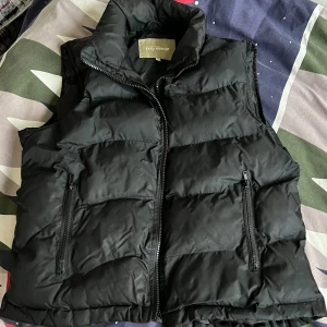 Svart dunväst från MyWear - Svart, quiltad dunväst från MyWear med hög krage och två dragkedjeförsedda fickor framtill. Västen har en klassisk puffer-look och stängs med dragkedja. Perfekt för lager-på-lager-stil under kyliga dagar.