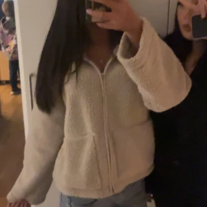 Beige teddyjacka med dragkedja - Mysig beige teddyjacka med dragkedja framtill och två stora fickor. Jackan har en avslappnad passform och är tillverkad i fluffigt fleece-material. Perfekt för kyliga dagar och enkel att matcha med jeans eller leggings.