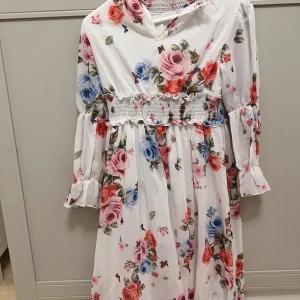 Vit blommig långklänning med volang - Romantisk långklänning i vitt med stora blommor i rosa, rött och blått. Klänningen har långärmade ärmar med volangdetaljer, smock i midjan och vid halsen samt en luftig, flowy passform. Perfekt för sommarens alla tillfällen.