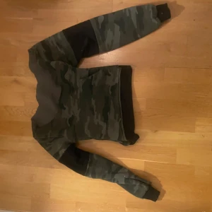 Camo-mönstrad sweatshirt i grönt - Säljer en grön camo-mönstrad sweatshirt med svarta muddar vid ärmslut och midja. 