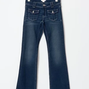 Mörkblå bootcut jeans med fickdetaljer - Snygga mörkblå jeans med bootcut passform och dekorativa fickor framtill med knappar. Jeansen har klassisk femficksdesign och subtila slitningar för en cool look. Byxorna är i storlek 170 passar om man vanligtvist har 36-38 och är runt 164 cm