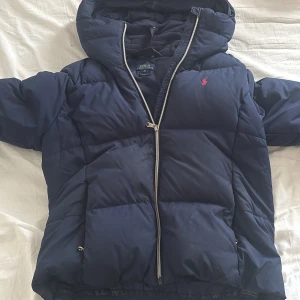 Ralph Lauren jacka !barn storlek! - Snygg mörkblå pufferjacka från Polo Ralph Lauren i barn storlek. Jackan har huva, dragkedja och den klassiska röda Polo-loggan på bröstet. Perfekt för kalla dagar med sin vadderade design och sportiga look. Xl barnstorlek