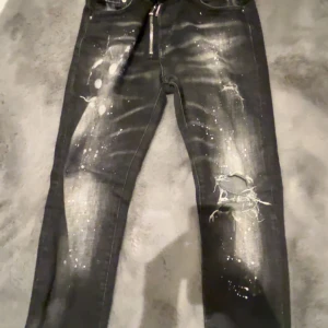 Svarta Dsquared2 jeans med färgstänk - Säljer ett par svarta Dsquared2 jeans i slim fit med slitningar och hål på knäet, samt coola vita färgstänk och dragkedja framtill. Jeansen har en snygg tvättad look och är tillverkade i Italien. Perfekta för dig som vill sticka ut med din stil.