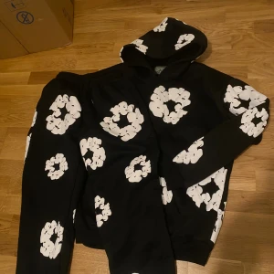 Denim Tears set svart hoodie och mjukis byxor med vita blommor - Svart hoodie och svart mjukis byxor från Denim Tears med stora vita blommönster över hela tröjan och byxorna. Tröjan har huva och långa ärmar, samt en avslappnad passform. Perfekt statement-plagg för dig som vill sticka ut med cool design.