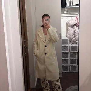 Beige kappa från Bershka - Snygg beige kappa från Bershka med klassisk krage och två svarta knappar framtill. Lång modell med raka ärmar och två fickor på sidorna. 