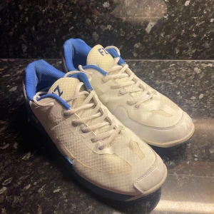 FZ Forza Badminton skor - Snygga sneakers från FZ Forza i vitt och blått med orange detaljer. Skorna har snörning, rund tå och platt sula. Ovandelen är i syntetmaterial med meshpaneler för extra andningsförmåga. Perfekta för dig som gillar sportig stil och vill sticka ut med färg. Skorna har lite slitningar på tårna och på sidan av skon, men det är inte som ger skorna sämre effekt