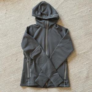 Grå hoodie från Houdini med dragkedja - Mörkgrå hoodie från Houdini med hel dragkedja framtill, två fickor med dragkedja och tumhål i ärmsluten. Tillverkad i mjuk fleeceblandning av polyester, polyamid och elastan. Perfekt för kyliga dagar och outdoor-aktiviteter.