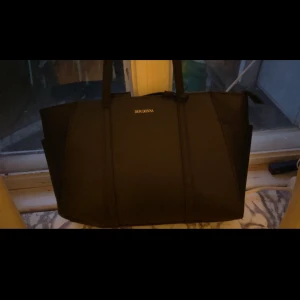 Svart väska från Don Donna - Stilren svart väska med gulddetaljer från Don Donna med två rejäla handtag och diskret logga i guld framtill. Väskan har en rymlig design och är perfekt för dig som vill ha plats med allt du behöver. Tillverkad i ett tåligt syntetmaterial.