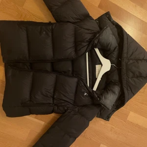 Svart pufferjacka Calvin Klein Jeans XS - Svart pufferjacka från Calvin Klein Jeans i storlek XS. Jackan har huva, dragkedja framtill och två sidofickor med dragkedja. Perfekt för kalla dagar och har en clean, stilren look som funkar till det mesta.