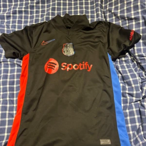 FC Barcelona svart matchtröja Nike - Säljer en svart FC Barcelona fotbollströja från Nike med röda och blå sidopaneler. Tröjan har klubbmärke, Spotify-logga och UNHCR-tryck. Klassisk V-ringning och korta ärmar, tillverkad i lätt och ventilerande material.