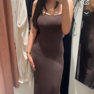 Brun långklänning  - Säljer en stilren brun långklänning med smala axelband och figurnära passform. Klänningen har en enkel design utan mönster och ärmlös siluett som framhäver kroppen. Perfekt för dig som gillar minimalistisk och trendig stil.