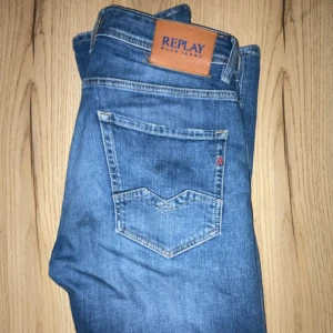 Blå Replay jeans med klassisk passform - Säljer ett par snygga blå jeans från Replay med klassisk femficksdesign och tydliga slitningar framtill. Jeansen har en normal midja och raka ben, med Replay-logga på bakfickan och läderpatch i midjan. Perfekt för dig som gillar stilrena och tidlösa jeans.