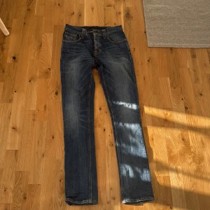 Nudie jeans grim trim - Snygga blå jeans från Nudie Jeans Co med klassisk femficksdesign och orangea kontrastsömmar. Jeansen har raka ben och coola slitningar på baksidan. Märkespatch i läder baktill och broderade detaljer på bakfickorna. Perfekt för dig som gillar stilrena jeans.