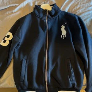 Polo Ralph Lauren Half Zip - Mörkblå tröja från Polo Ralph Lauren med stor vit logga på bröstet och siffra 3 på ärmen. Tröjan har dragkedja framtill, ribbade muddar med vita ränder och två fickor. Tillverkad i mjuk bomull för en skön och sportig look. Mycket bra skick. Storlek S.