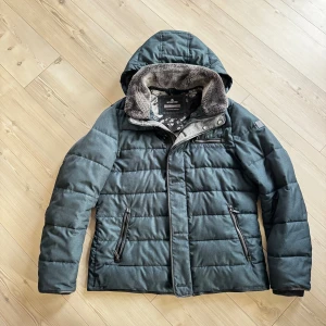 Exklusiv Mörkgrön pufferjacka från Milestone - Snygg mörkgrön pufferjacka från Milestone med huva och mjukt foder i kragen. Jackan har quiltad design, dragkedjor på fickorna och en extra bröstficka med dragkedja. Perfekt för kalla dagar med sin varma och fluffiga känsla.