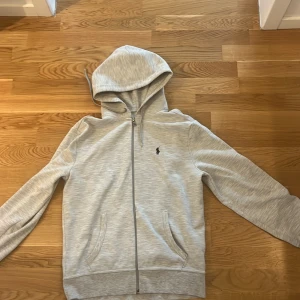 Ljusgrå hoodie från Polo Ralph Lauren - Snygg ljusgrå hoodie från Polo Ralph Lauren med dragkedja och klassisk svart logga på bröstet. Tröjan har huva med snörning, två fickor framtill och långa ärmar. Perfekt för en chill och stilren look.