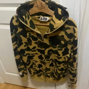 BAPE camo hoodie med dragkedja M - Säljer en ikonisk camo hoodie från BAPE i grönt, svart och beige. Tröjan har hel dragkedja, broderade detaljer på huvan och klassisk BAPE shark-design. Tillverkad i bomull och har känguruficka framtill. Perfekt för dig som vill sticka ut med streetwear.