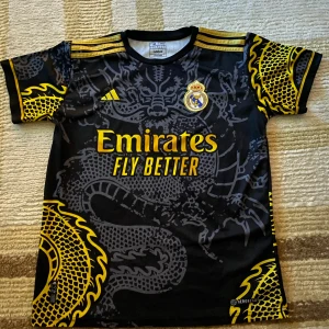 Real Madrid Ronaldo 7 svart/gul tröja - Säljer en Real Madrid fotbollströja från Adidas med Ronaldo 7 på ryggen. Tröjan är svart med stora drakmönster i grått och gult, samt gula detaljer och tryck. Kortärmad modell i lätt funktionsmaterial, perfekt för match eller träning.