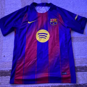 Barcelona FC kortärmad matchtröja Nike - Säljer en officiell Barcelona FC fotbollströja från Nike med klassiska blå och röda vertikala ränder. Tröjan har klubbmärke, gul Nike-logga och Spotify-sponsor på framsidan samt UNHCR-tryck på ryggen. Tillverkad i lätt och ventilerande polyester, perfekt för träning eller match.
