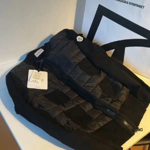 Svart dunväst från Moncler, storlek S - Svart dunväst från Moncler med quiltad front och stickade sidor. Västen har dragkedja framtill och klassisk Moncler-logga på axeln. Materialmix av dun och stickad polyester ger en snygg och modern look. Perfekt för lager-på-lager och streetstyle.