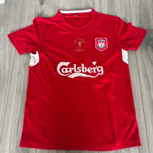Liverpool FC 2005 Champions tröja S - Liverpool FC fotbollströja i rött med vita detaljer och broderat klubbmärke samt guldtext 'European Champions Istanbul 2005'. Carlsberg-logga på bröstet. Klassisk passform i funktionsmaterial, perfekt för fans.
