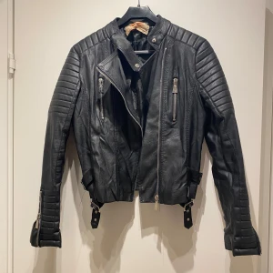 Svart skinnjacka Toyelles med detaljer - Cool svart skinnjacka från Toyelles med quiltade axlar och armar, flera dragkedjor och spännen i midjan. Jackan har lång ärm och snygga detaljer. Har lite märken på axlarna annars bra skick (se bilderna)