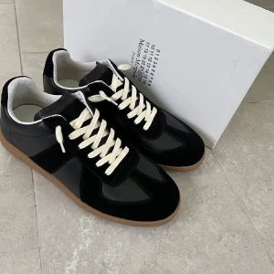 Maison Margiela svarta sneakers - Maison Margiela sneakers i svart skinn och mocka med kontrasterande vita snören och gummisula i beige. Klassisk rund tå och platt sula, stilren design med diskreta detaljer. Perfekt för dig som gillar minimalistisk och lyxig streetwear.
