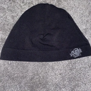 Svart mössa från SYNA world - Svart mössa från SYNA med stilren, böjd form och en grå logga i graffiti-stil framtill. Tillverkad i mjuk polyester och elastan som sitter skönt på huvudet. Perfekt för dig som gillar streetwear och vill ha en enkel men snygg accessoar.har ej påsen