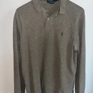 Grå långärmad pikétröja Ralph Lauren - Snygg grå långärmad pikétröja från Ralph Lauren med klassisk krage och knäppning framtill. Tröjan har den ikoniska gröna broderade loggan på bröstet och är tillverkad i mjuk bomull. Perfekt för en clean och stilren look.