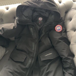 Svart dunjacka från Canada Goose L - Säljer en svart dunjacka från Canada Goose med huva och klassisk logga på ärmen. Jackan har flera stora fickor framtill, dragkedja och är perfekt för kalla dagar. Stilen är clean och tidlös, passar dig som vill ha både värme och streetkänsla.