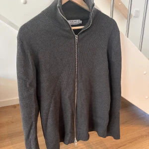 Mörkgrå ribbad kofta från Ljung - Säljer en stilren mörkgrå kofta från Ljung i storlek M. Koftan är ribbstickad med hög krage och har en hel dragkedja framtill. Materialet är mjukt och stickat, perfekt för kyliga dagar. Passformen är normal och ärmarna är långa.