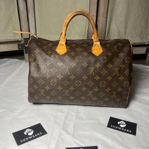 Louis Vuitton Speedy 30 - Klassisk Louis Vuitton Speedy handväska i brunt monogrammönster med ikoniska LV-loggor. Väskan har rund form, två robusta handtag i ljust skinn och dragkedja upptill. Tillverkad i canvas med detaljer i skinn och guldiga metalldelar. Perfekt för dig som gillar lyxig och tidlös stil.