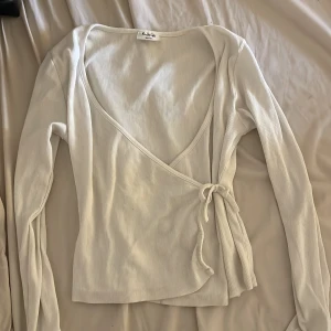 Vit omlottopp från Kappahl - Säljer en vit omlottopp från Kappahl i ribbad bomull. Toppen har långa ärmar, v-ringad hals och knyt i sidan för en snygg passform. Perfekt att styla med jeans eller kjol för en fräsch look.