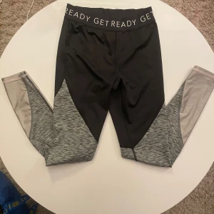 Svarta och grå träningsleggings H&M - Snygga träningsleggings från H&M Sport med svart överdel och gråmelerade partier på benen. Elastisk midja med texten 'GET READY' och inslag av ljusrosa vid bensluten. Perfekta för gymmet eller löprundan.