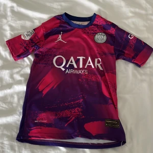 PSG Mbappé fotbollströja Jordan 164 - Snygg PSG fotbollströja med Mbappé 7 på ryggen, tillverkad i lätt och ventilerande material. Tröjan har coolt röd-lila mönster, vita tryck och Jordan-logga på bröstet. Perfekt för dig som älskar fotboll och vill sticka ut på planen.