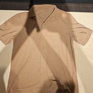 Beige manchester kortärmad skjorta - Kortärmad skjorta i beige manchester med diskreta vertikala ränder. Skjortan har klassisk krage och knäppning framtill med fyra knappar. Perfekt för dig som gillar retrovibbar och mjuka material.