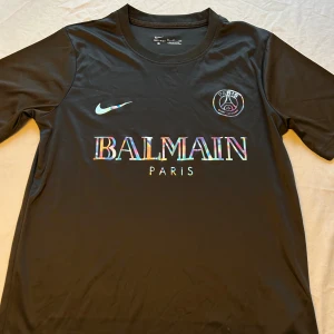 Svart PSG x Balmain fotbollströja Nike - Snygg svart fotbollströja  Nike i samarbete med PSG och Balmain. Tröjan har korta ärmar, rund hals och är gjord i lätt, andasvänligt Dri-FIT-material. Framsidan pryds av holografiska loggor och text. Perfekt för dig som vill sticka ut på planen.