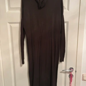 Brun långärmad klänning från JCL - Mörkbrun långklänning från JCL med lång ärm och enkel, rak passform. Klänningen är tillverkad i ett mjukt, stickat material som känns skönt mot huden. Perfekt för dig som gillar minimalistisk och stilren look.