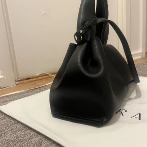 Svart handväska från Swedora i skinn - Stilren svart handväska från Swedora i mjukt skinn med breda handtag och justerbar axelrem. Väskan har minimalistisk design med guldfärgade metalldetaljer och levereras i en vit dustbag med logga. Perfekt för dig som gillar klassisk och clean look.