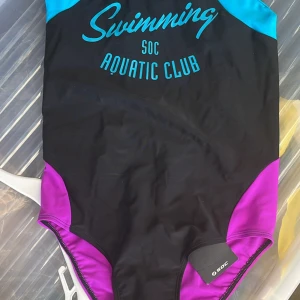 Svart baddräkt från SOC med lila detaljer till flicka 158/164 - Snygg svart baddräkt från SOC med lila och turkosa detaljer. Tryck med texten 'Swimming SOC Aquatic Club' framtill. Perfekt för dig som gillar att simma och vill ha en sportig look vid poolen eller stranden.