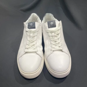 Vita sneakers från GANT i skinn - Fräscha vita sneakers från GANT i skinn med klassisk design. Skorna har vit ovandel, vit sula och mörkblå detaljer på häl och plös. Snygg GANT-logga på sidan och plösen. Perfekta för en clean och stilren look. Nypris 1800-2000