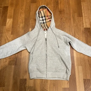 Grå hoodie från Burberry med dragkedja - Snygg grå hoodie från Burberry med klassiskt rutigt foder i huvan och broderad logga på bröstet. Tröjan har dragkedja framtill, snörning i huvan och två fickor. Perfekt för dig som vill ha en stilren och lyxig look.