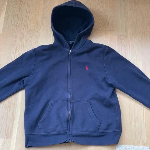Mörkblå hoodie från Polo Ralph Lauren - Mörkblå hoodie från Polo Ralph Lauren med dragkedja framtill och klassisk röd logga på bröstet. Tröjan har huva, känguruficka och är tillverkad i mjuk bomullsmix. Perfekt för chill dagar eller när du vill ha en clean och stilren look.