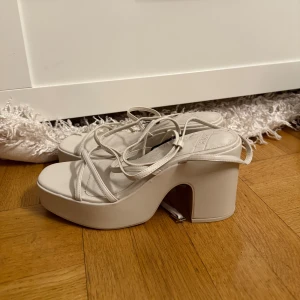Vita platåsandaler med klack från Zara - Säljer ett par trendiga vita sandaler från Zara med höga platåklackar och tunna remmar som snöras runt foten. Skorna har öppen tå och en chunky klack som ger extra höjd. Perfekta för dig som vill sticka ut med en cool och modern look.