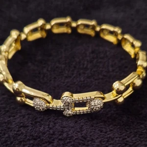 Förgyllt armband med detaljer av zirconia pavé hästsko - Unikt armband i guldton med länkar som har rundade och rektangulära hästskoformer. Vissa länkar är dekorerade med små zirconia stenar insatta med klor i pavé- teknik som ger extra bling. Armbandet är öppningsbart och ser ut som mycket dyrare än bijouteri p.g. a perfekt utförande och steninsättning.Designen är modern och sticker ut med sina geometriska former och skimrande detaljer. Perfekt för dig som gillar att synas.